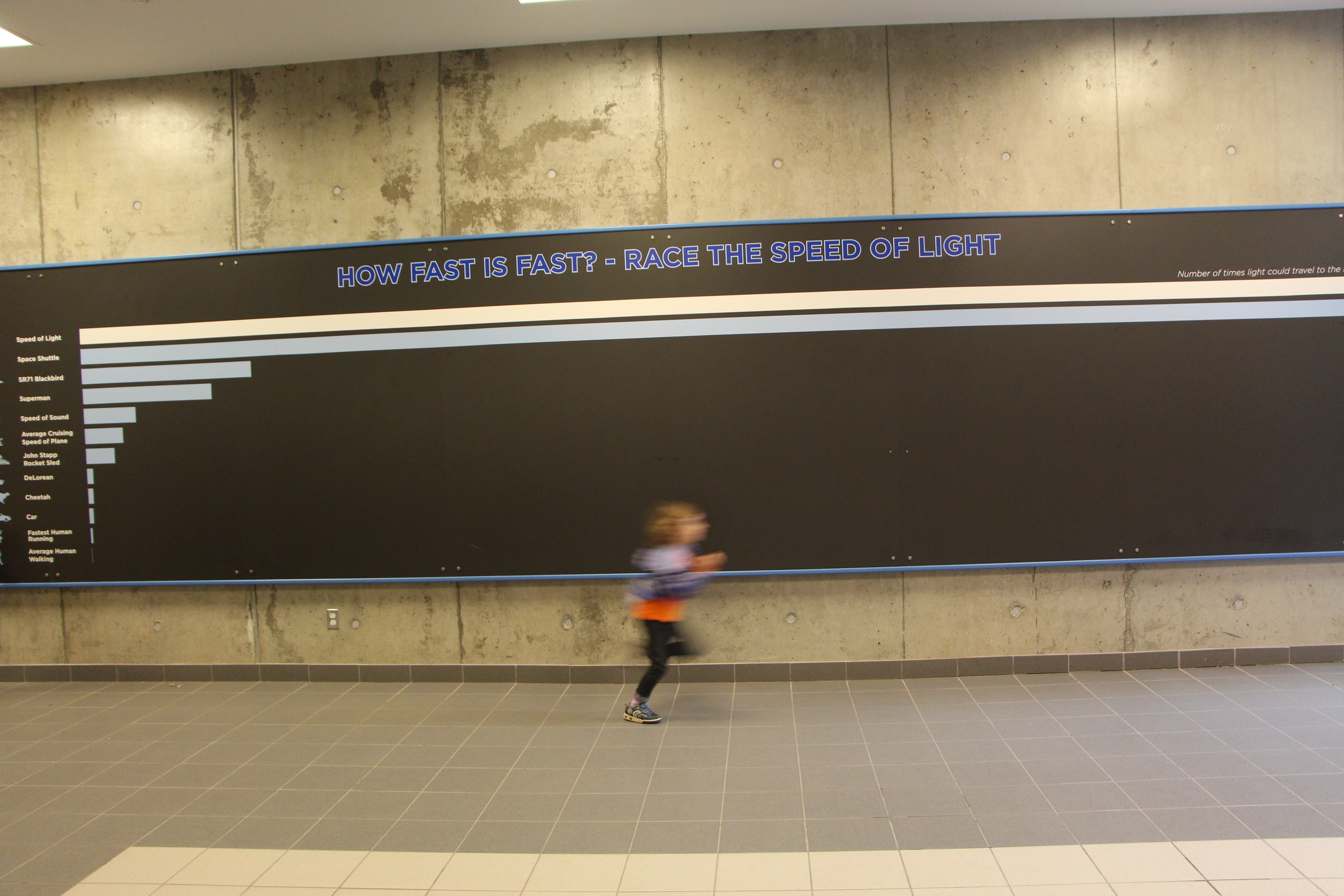 A small child running in front of a "race the speed of light" exhibit