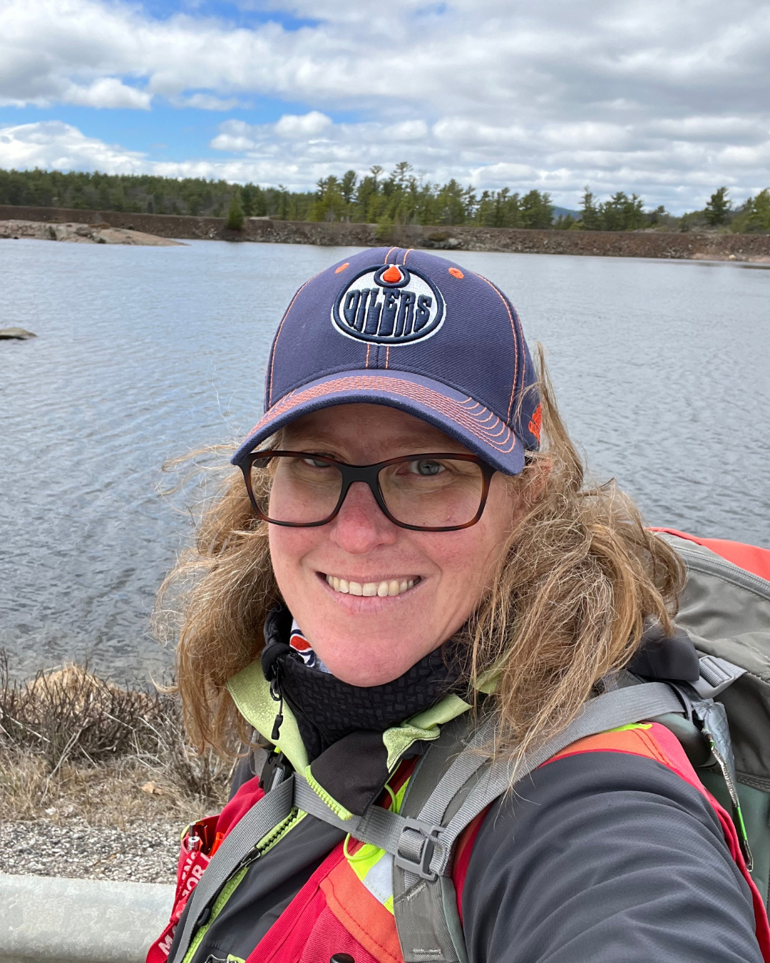 Jen is wearing an Oilers baseball cap, a rain jacket and a backpack. She is standing in front of a lake. 