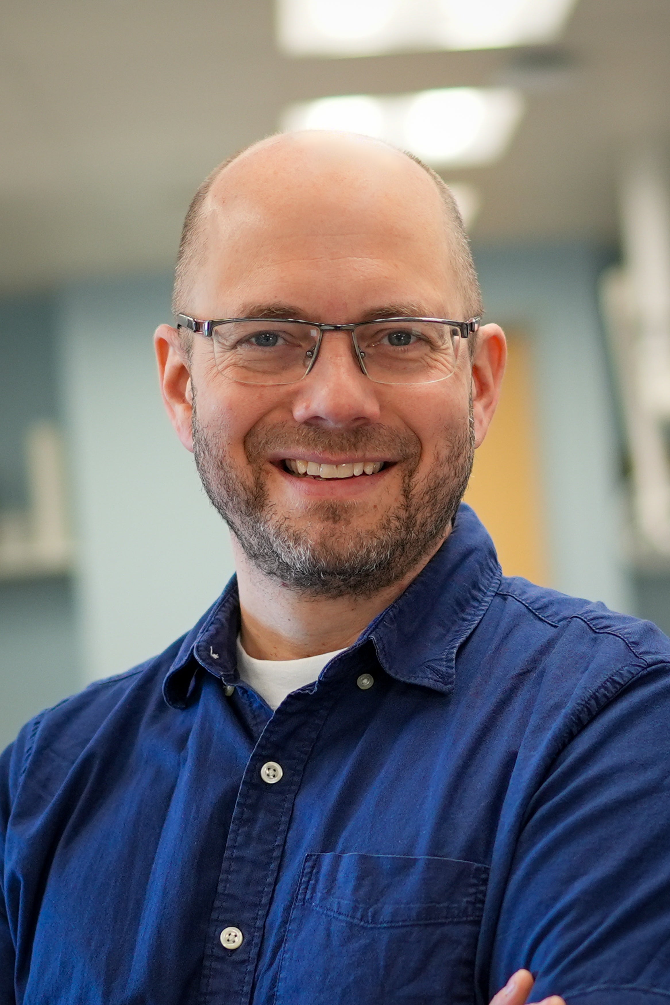 Head-and-shoulders portrait of Josh Neufeld a person wearing glasses and a blue button-up shirt, facing the camera in an indoor hallway with soft overhead lighting and a blurred background.