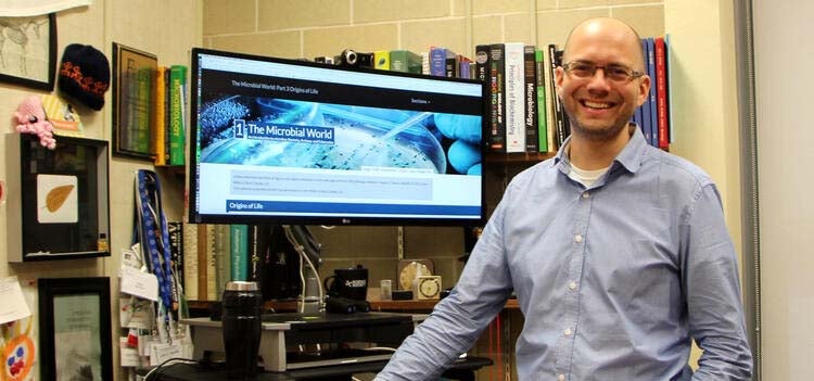 Josh Neufeld standing beside a desktop displaying the Microbiology 240 course homepage.