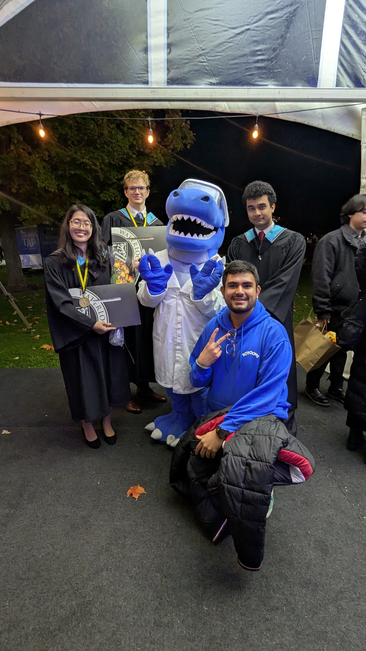Four graduates dressed in their convocation gowns and hoods posing with Cobalt the mascot