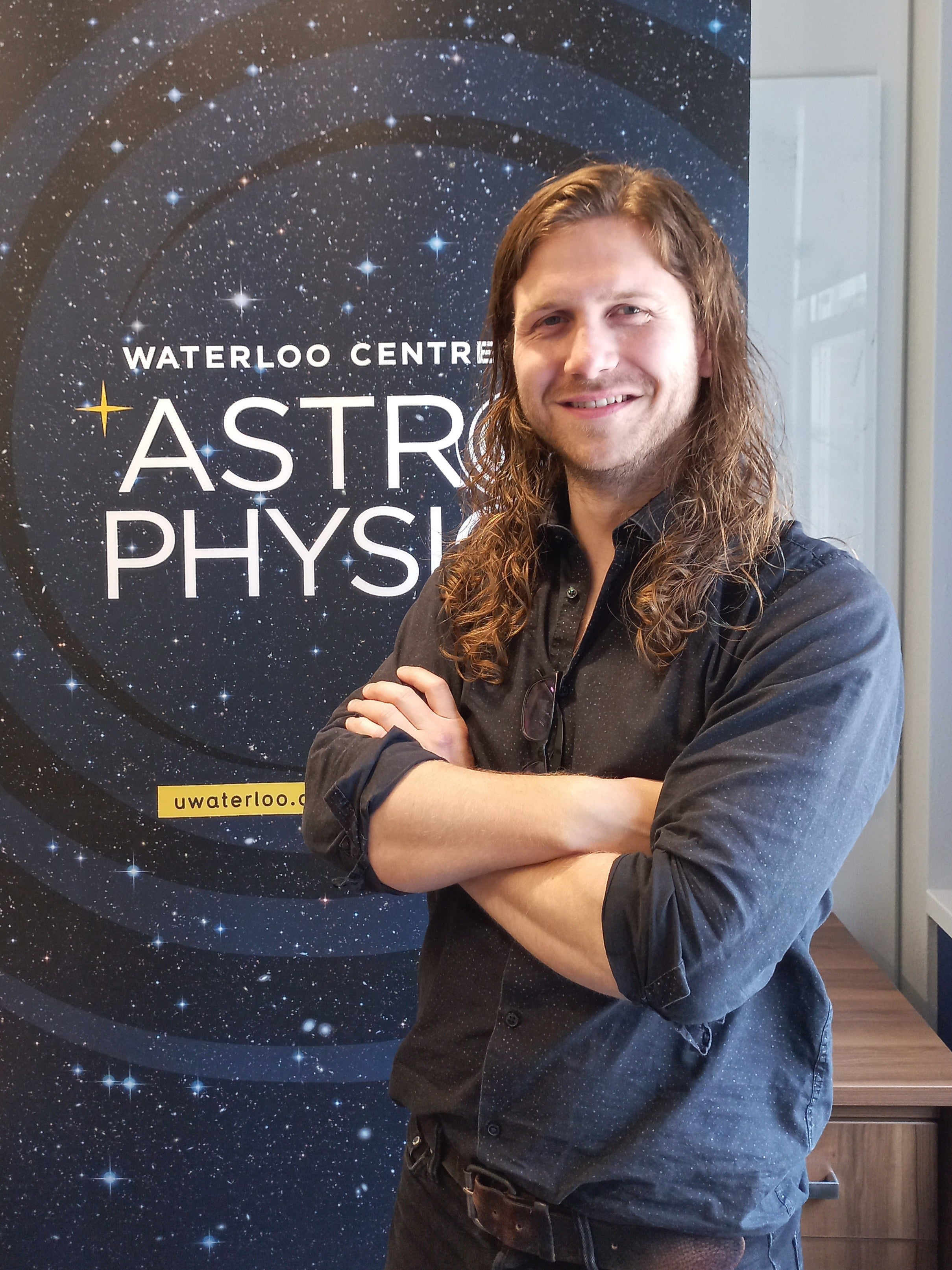 Roan Haggar (he/him) is standing in front of a banner that says "Waterloo Centre for Astro Physics."