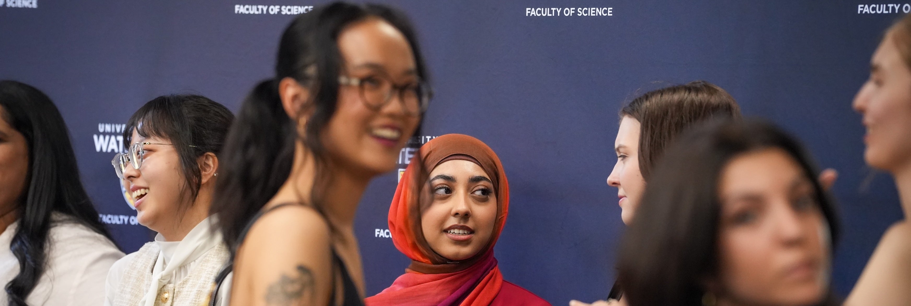 A group of female students standing in front of a University of Waterloo logo backdrop. 