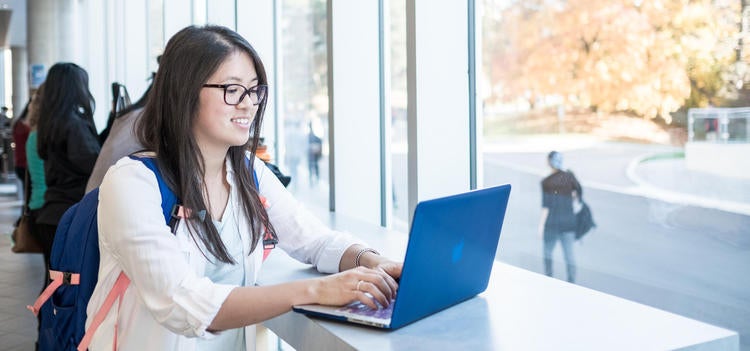 Asian female student typing on blue laptop by a window.