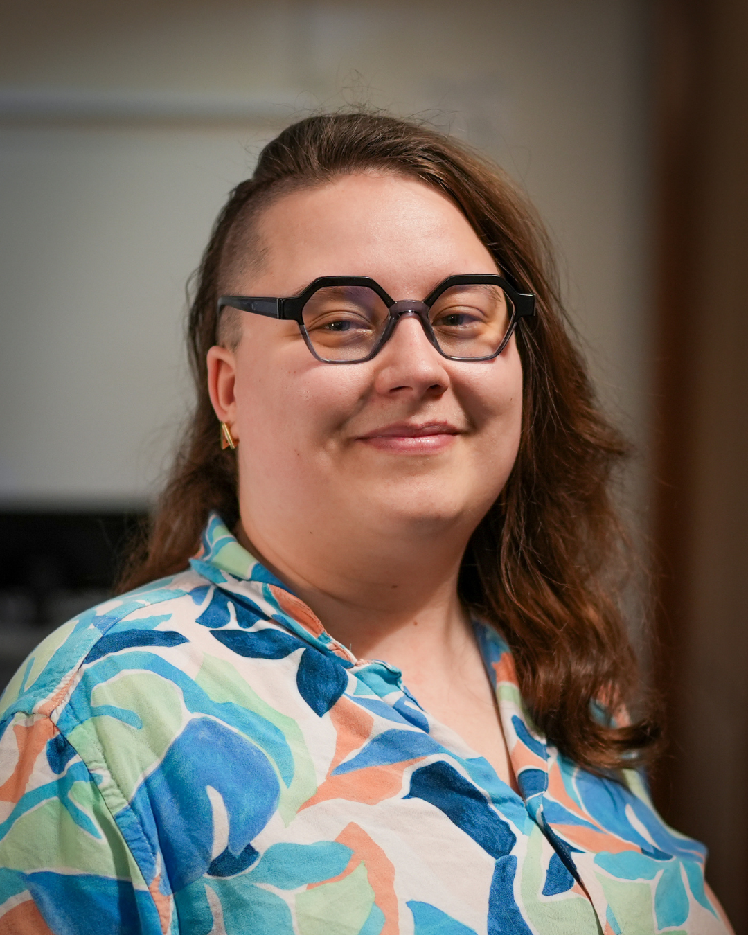 A headshot of Taylor. She has brown hair and is wearing glasses and a blue patterned collared shirt. 