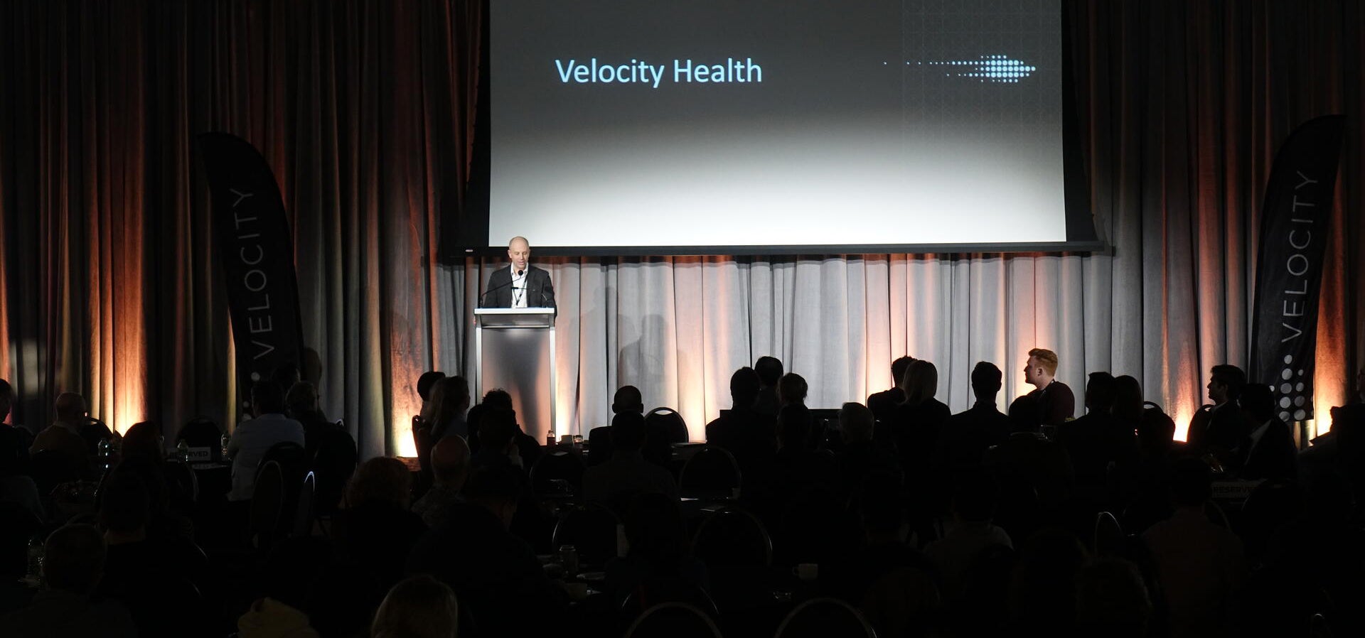 Man speaking at podium with Velocity Health on screen behind him.