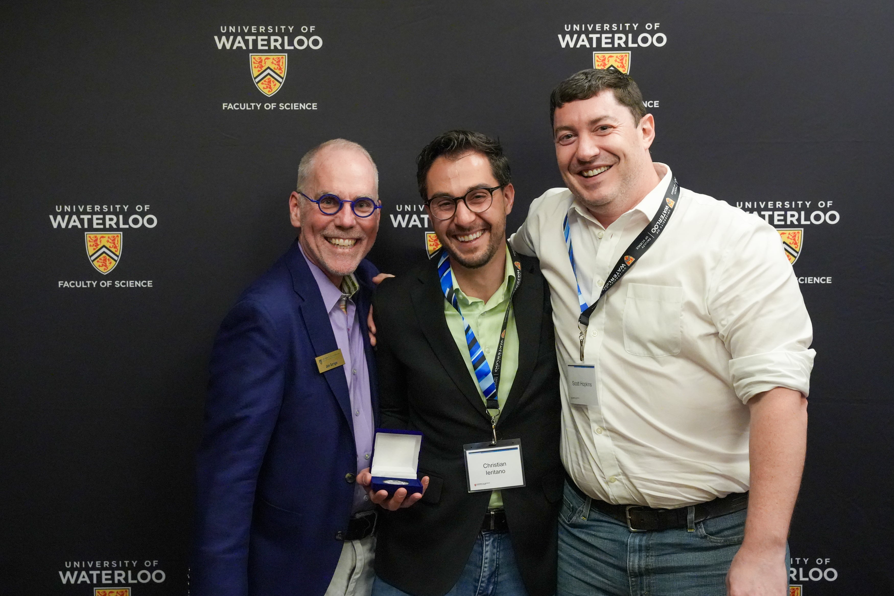 Three men standing against a black background printed with the University of Waterloo logo. One man is holding a medal.
