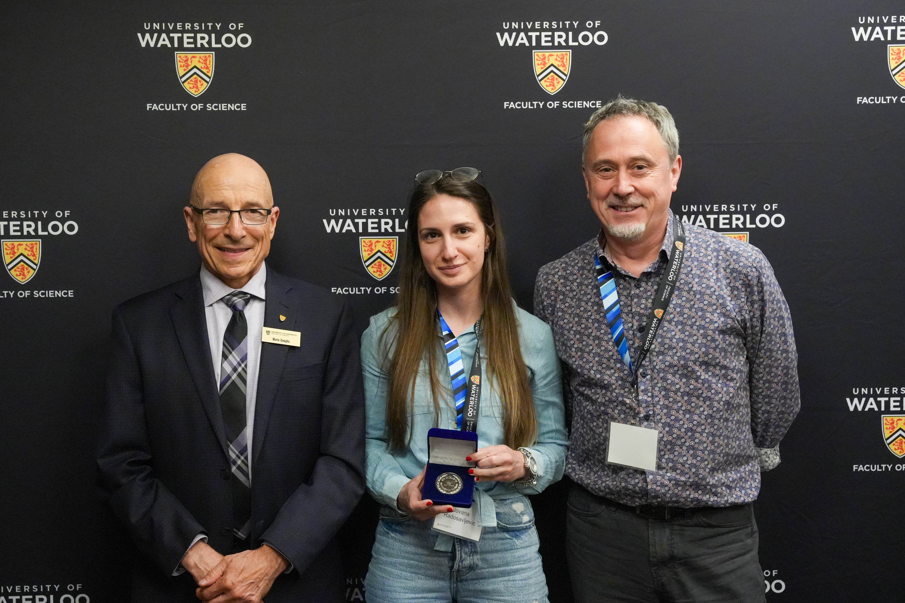 Two men and a woman standing against a black background printed with the University of Waterloo logo. The woman is holding a medal.