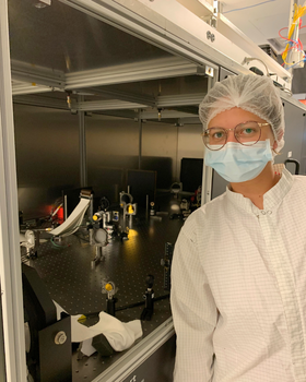 wearing a cleanroom suit, hair net, face mask, and glasses stands beside an experiment setup with mirrors, lenses, and precision instruments on a metal table inside a laboratory.