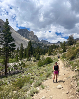 Ariana hiking along a dirt trail surrounded by alpine scenery with pine trees, rocky terrain, and tall mountain peaks in the background under a partly cloudy sky.