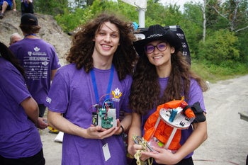 Two smiling students wearing matching purple Waterloo Rocketry T-shirts stand outdoors, posing together while holding electronic rocket components.