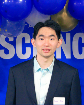 Daniel Wang has short dark hair. He is wearing a light blue collared shirt with a navy blue blazer over top. There is a blue background with balloons surrounding him.