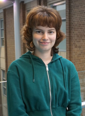 Grace has short brown hair and is smiling. She is wearing a green hoodie. There is a brown brick wall in the background.