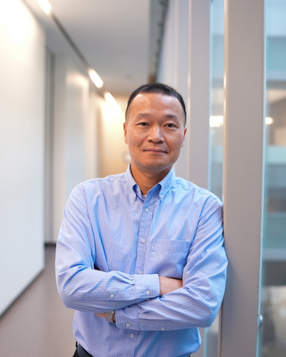 Juewen Liu leaning against a wall in a long hallway. His arms are crossed and he his wearing a blue button down dress shirt.