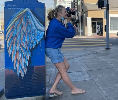 Olga standing with her back against a painting of wings, with a camera to her eye
