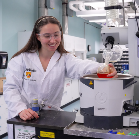 A female student conducting a chemistry student wearing a white lab coat and goggles,