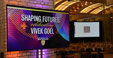 Digital sign reading "Shaping Futures: Celebrating Vivek Goel" in a large room set up with tables set for dinner