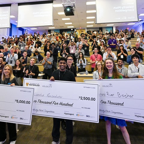 A room full of students with three students in the front holding big money cheques
