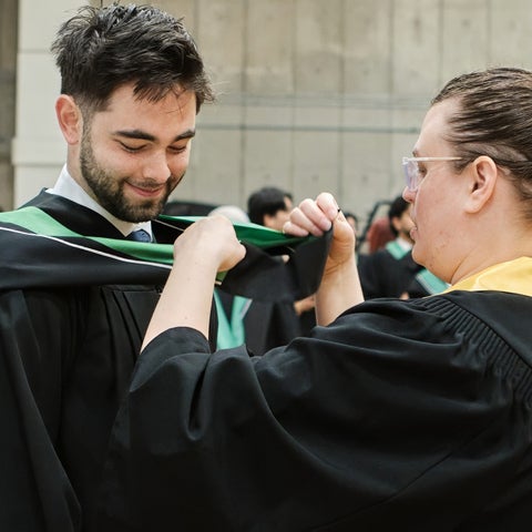 University of Waterloo staff in volunteer regalia helping a Faculty of Arts graduand fix their faculty hood