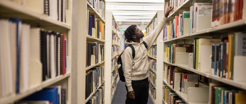 Waterloo student wearing hoodie and backpack in the middle of a Library aisle picking out a book on the top shelf