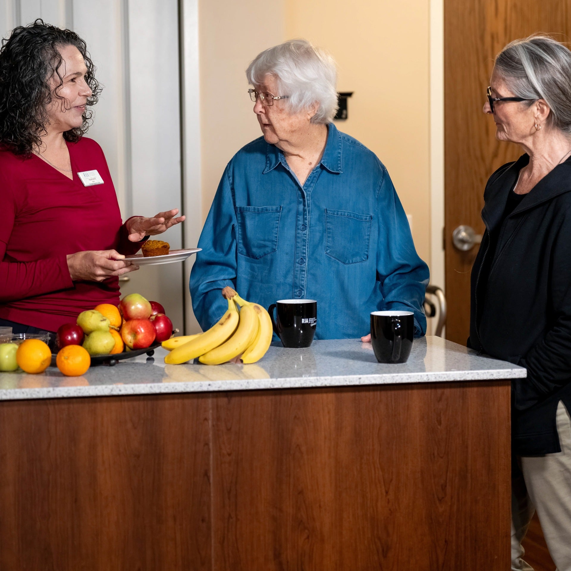 Heather Keller chatting with two senior residents while holding a muffin inside a kitchen with fruits and mugs on top of a counter 