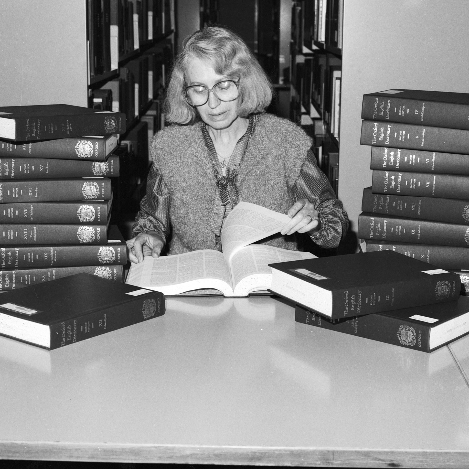 Woman reading Oxford English Dictionary at the library in between stack of dictionaries on the table