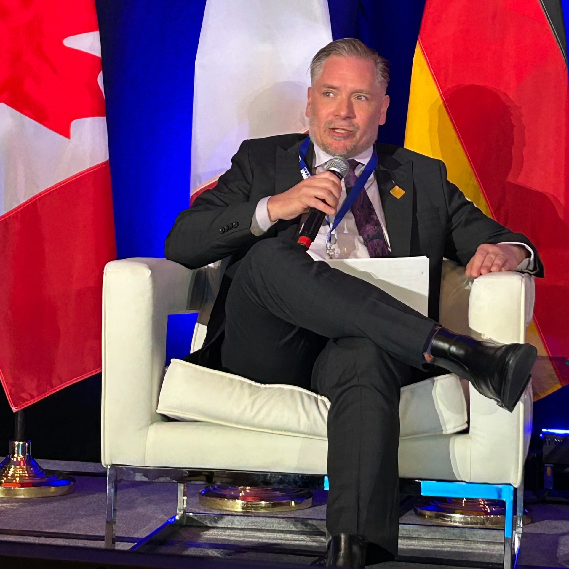 Mark Schaan speaking on stage sitting on a chair in front of Canadian flags