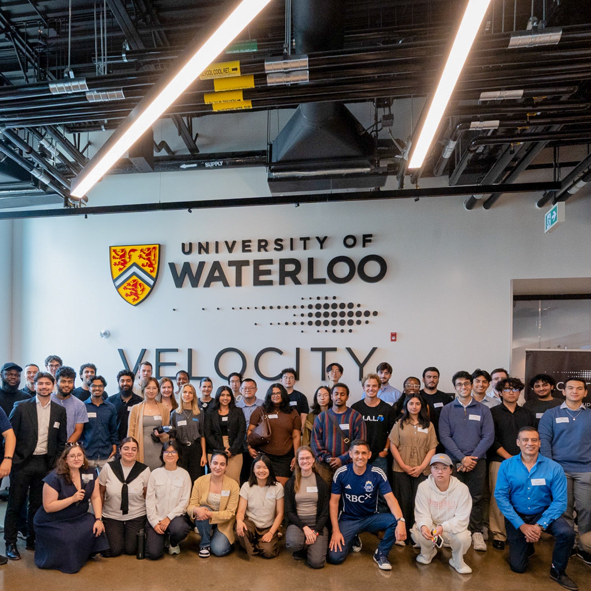 Cohort of the Summer Accelerator program posing together in front of the University of Waterloo and Velocity sign inside the Innovation Arena