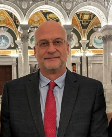 Christian Smith, wearing a blue shirt, red tie, and dark suit jacket, smiles in front of a background of mosaics and columns.