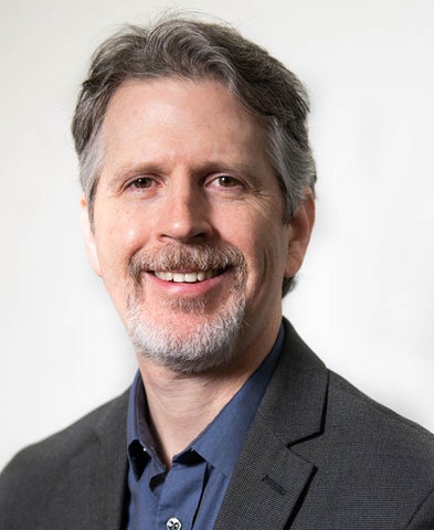 Gary J. Adler Jr. smiling in a suit jacket and blue shirt in front of a white background