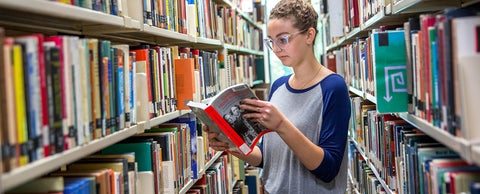 A student looks at a book while standing in between shelves of books in the St. Jerome's University Library