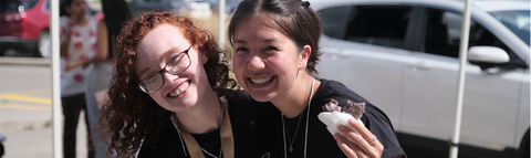 Two female students with their arms around each other smiling - one is eating a donut