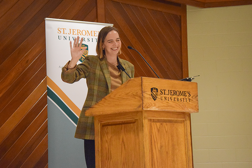 Colleen Dulle waving hello to the audience from the podium at St. Jerome's University Chapel in front of a standing banner that shows the St. Jerome's logo