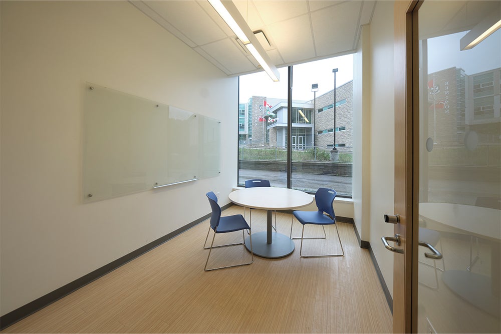 Brightly lit residence study room with whiteboard, table, chairs and large window overlooking the campus