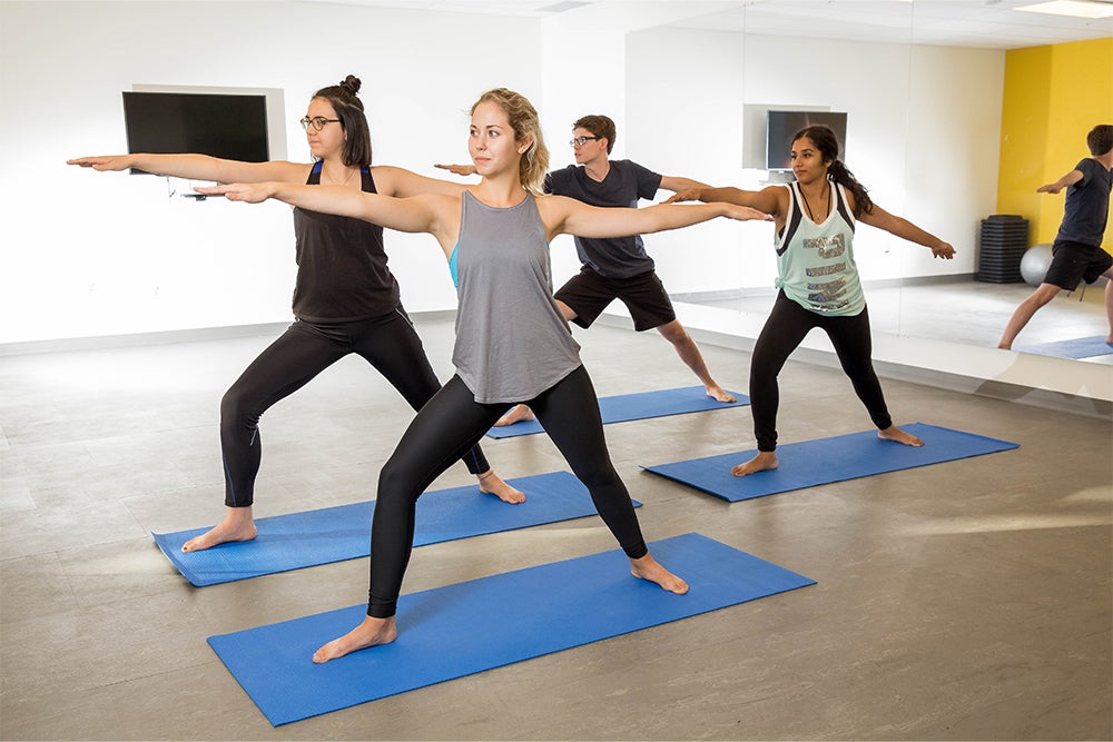 Students stand in warrior yoga pose on yoga mats in the fitness room in Siegfriend and Ryan Hall