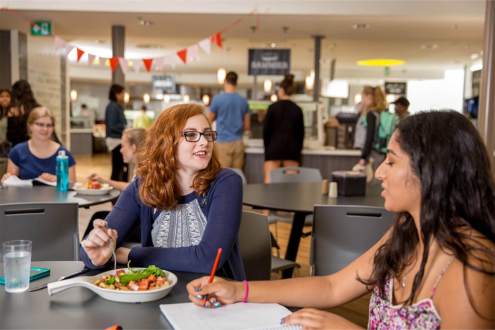 Two students writing in books and eat heaping plates of all-you-can-eat cafeteria food 