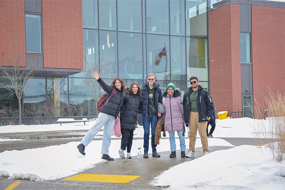 Five students dressed in winter coats, hats and mitts stand together in from of the St. Jerome's Atrium entrance