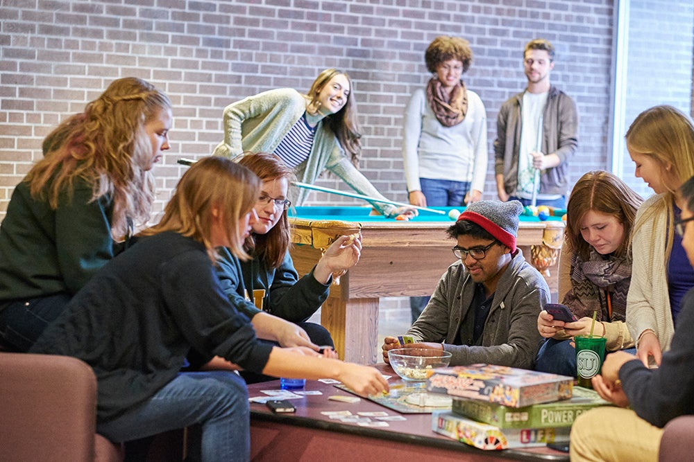 Students sit together playing games at a table while others play at a pool table behind them in the St. Jerome's Fireplace lounge