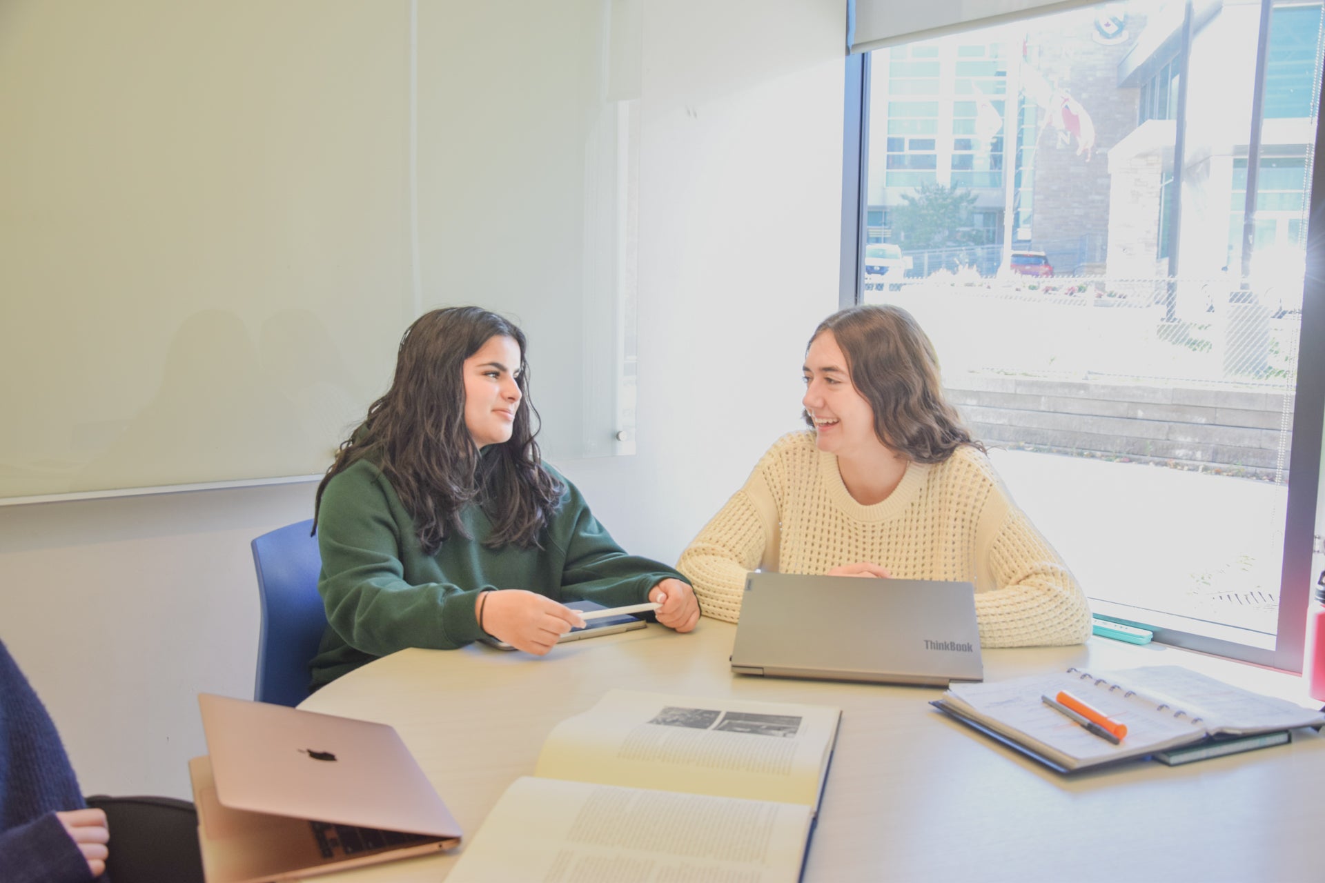 Two students study at a table and chairs beside a window overlooking the campus in a clean, bright study room