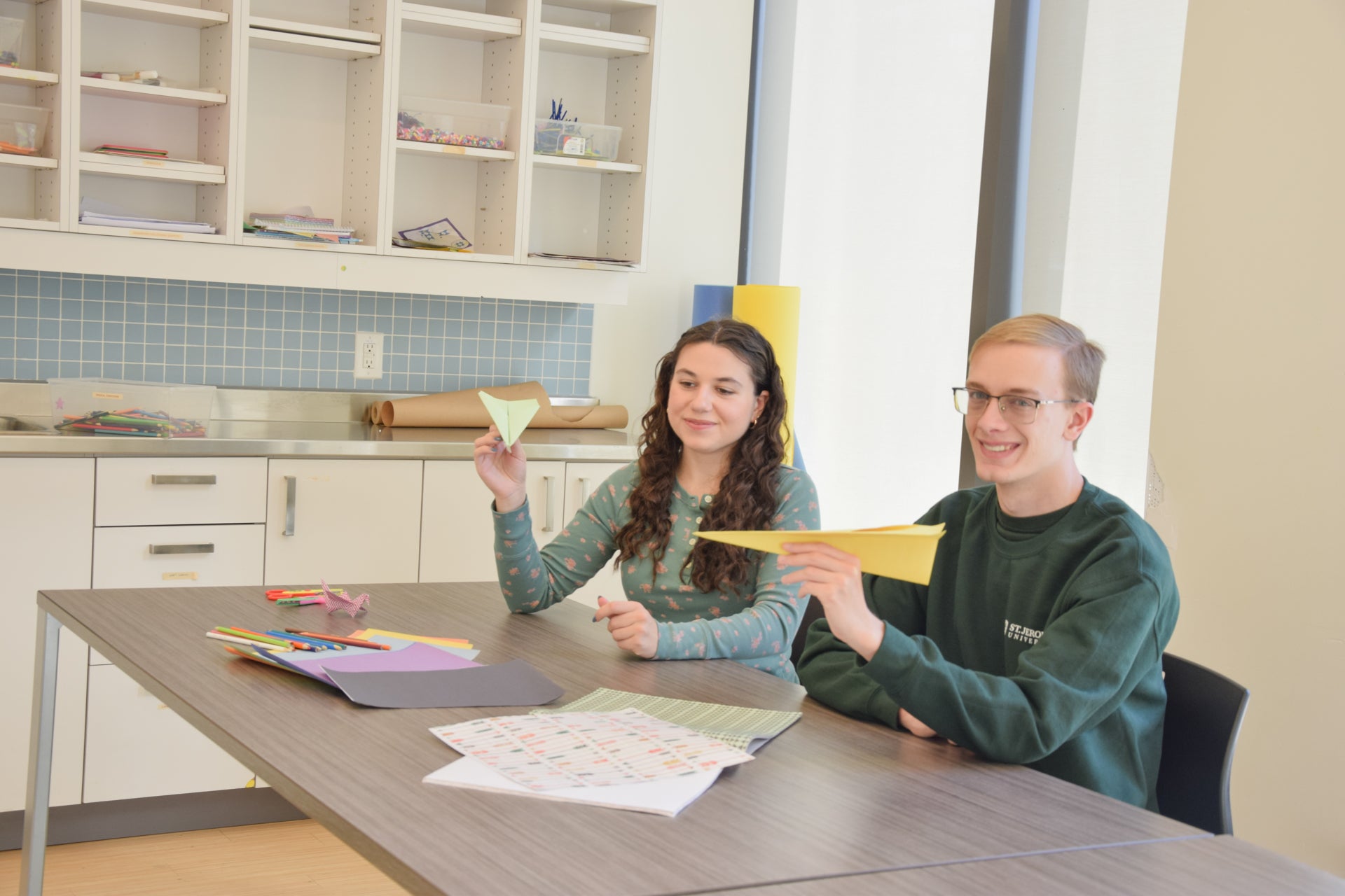 Two students showing paper airplanes they made with the brightly coloured paper supplied in the clean, spacious craft room with sun shining through windows overlooking the campus