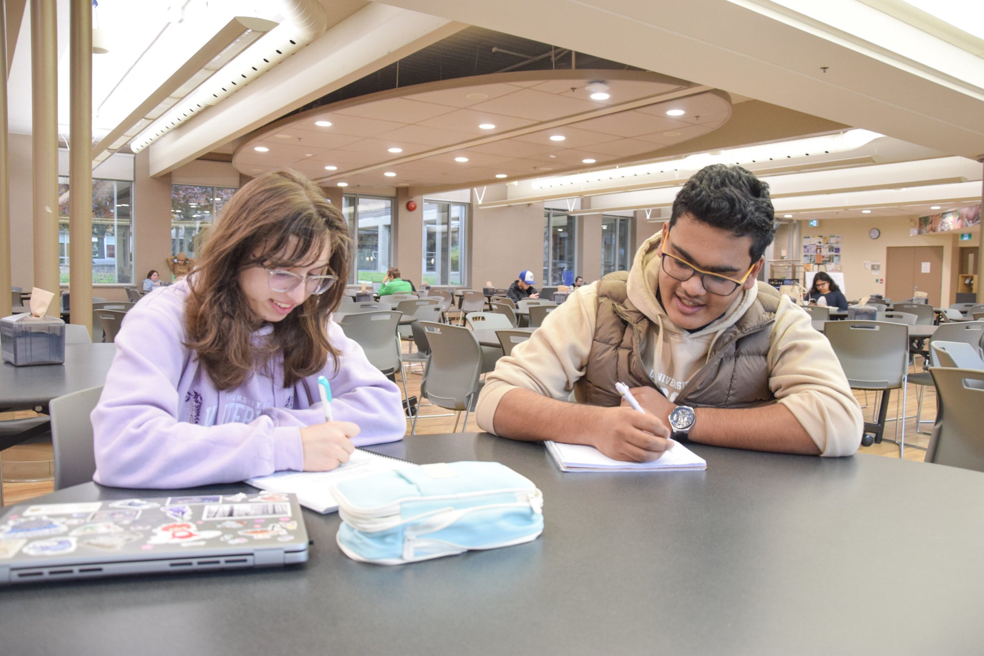 two students writing in notebooks at one of the many tables in the Doug Letson Community Center