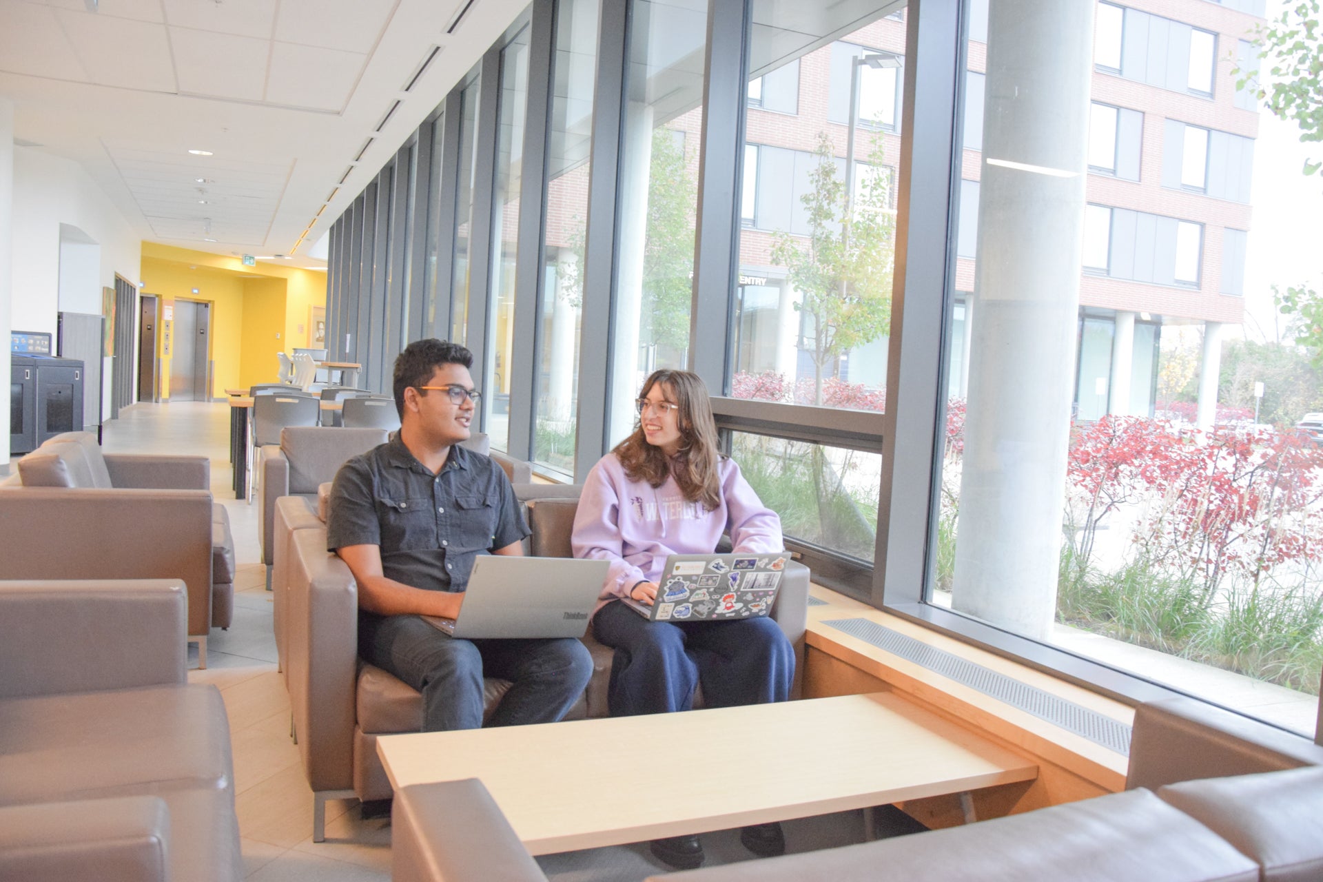 Two students study in cushioned lounge chairs, large windows overlooking the residence parking lot and trees with bright red autumn leaves 