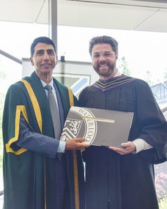 Andrew Zettel smiles and holds his diploma, on his left is Bruce Rodrigues, SJU Chancellor