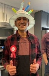 Anu Khanna smiles and gives a double thumbs up wearing a birthday cake hat.