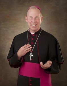 Most Reverend Gary M. Gordon stands in front of a brown background in bishop's robes, smiling and holding a hand out