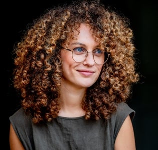 Maya Venters smiles in front of a black background in a grey shirt with an abundance of curly hair