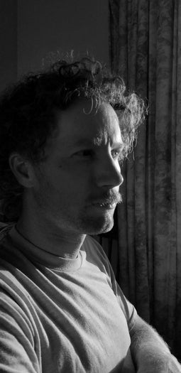 Nicholas Herring in side profile looks off in the distance pensively against a pain background in black and white.