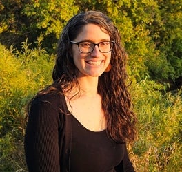 Rebecca Mancini smiles in from of a sunlit background of trees and nature