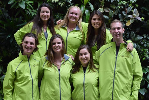Seven members of the St. Paul's Don team pose with green jackets in front of a living wall