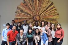 A group of fifteen students pose in front of wooden sunburst wall art